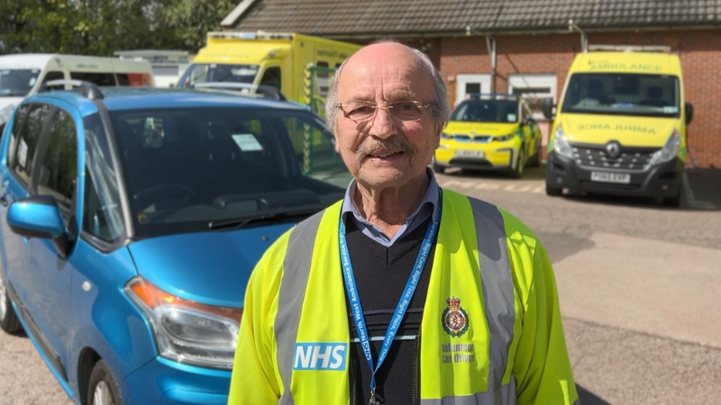 Volunteer Car Driver Colin standing in front of his car in his uniform smiling.